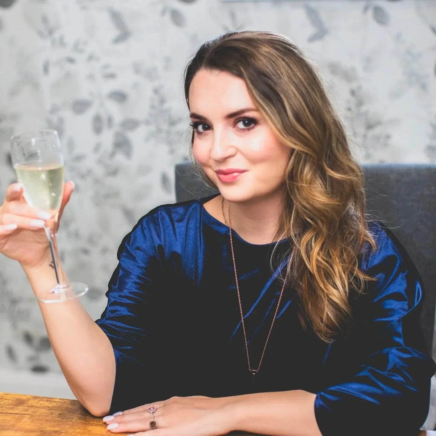 Willow Winters sits at a table, wearing a blue shirt, holding a glass of champagne.