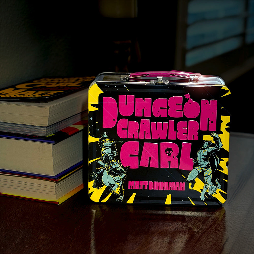 A metal lunchbox titled "Dungeon Crawler Carl" by Matt Dinniman, shown standing on a dark wooden surface next to stacked books. The design is dominated by black, bright pink, and yellow, with illustrated characters and comic-style text.