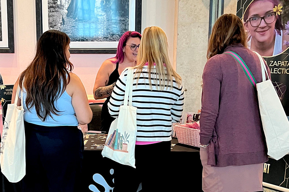 Three women holding tote bags, looking at the merchandise on the Ink Games Publishing table at the Romantically Yours book event.