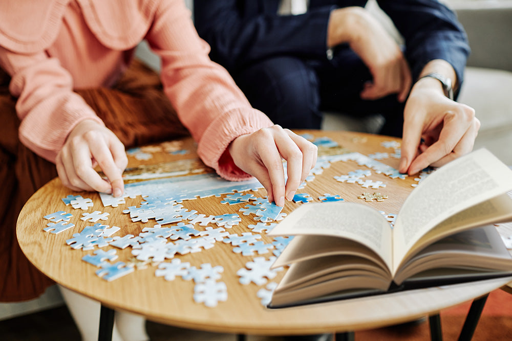 Two people working on a puzzle together with an open book on a table.